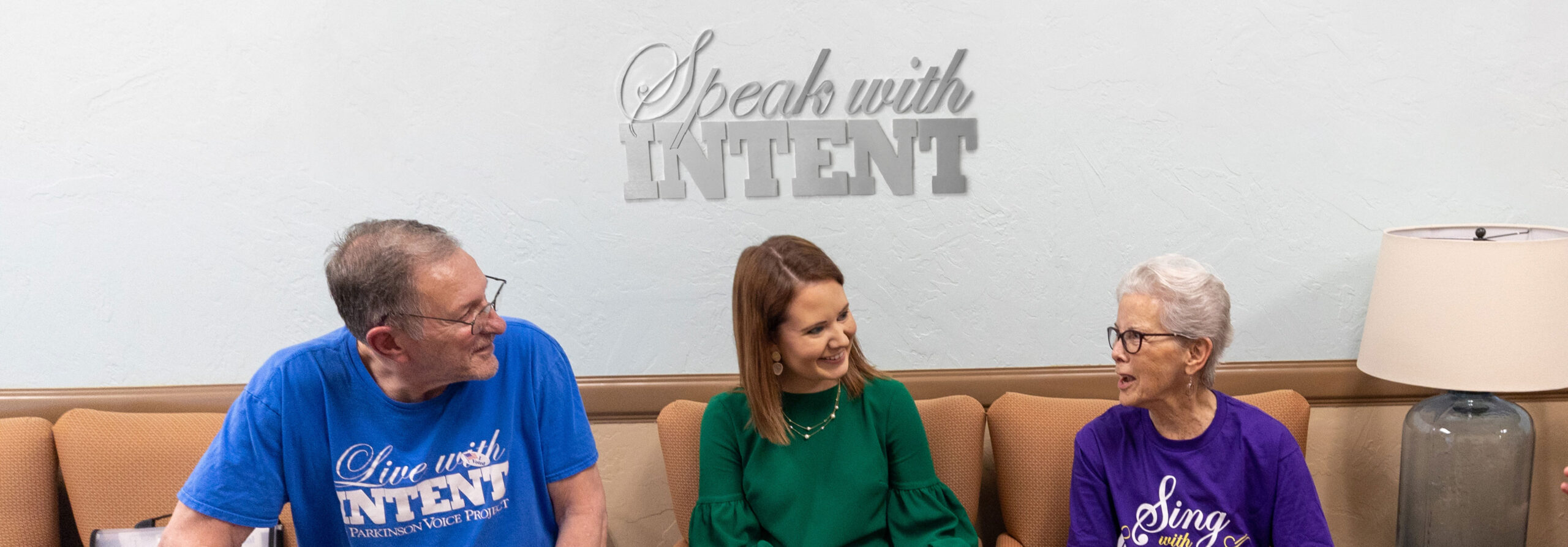 Three people talking in the Parkinson Voice Project headquarters under a sign that says "Speak with INTENT"